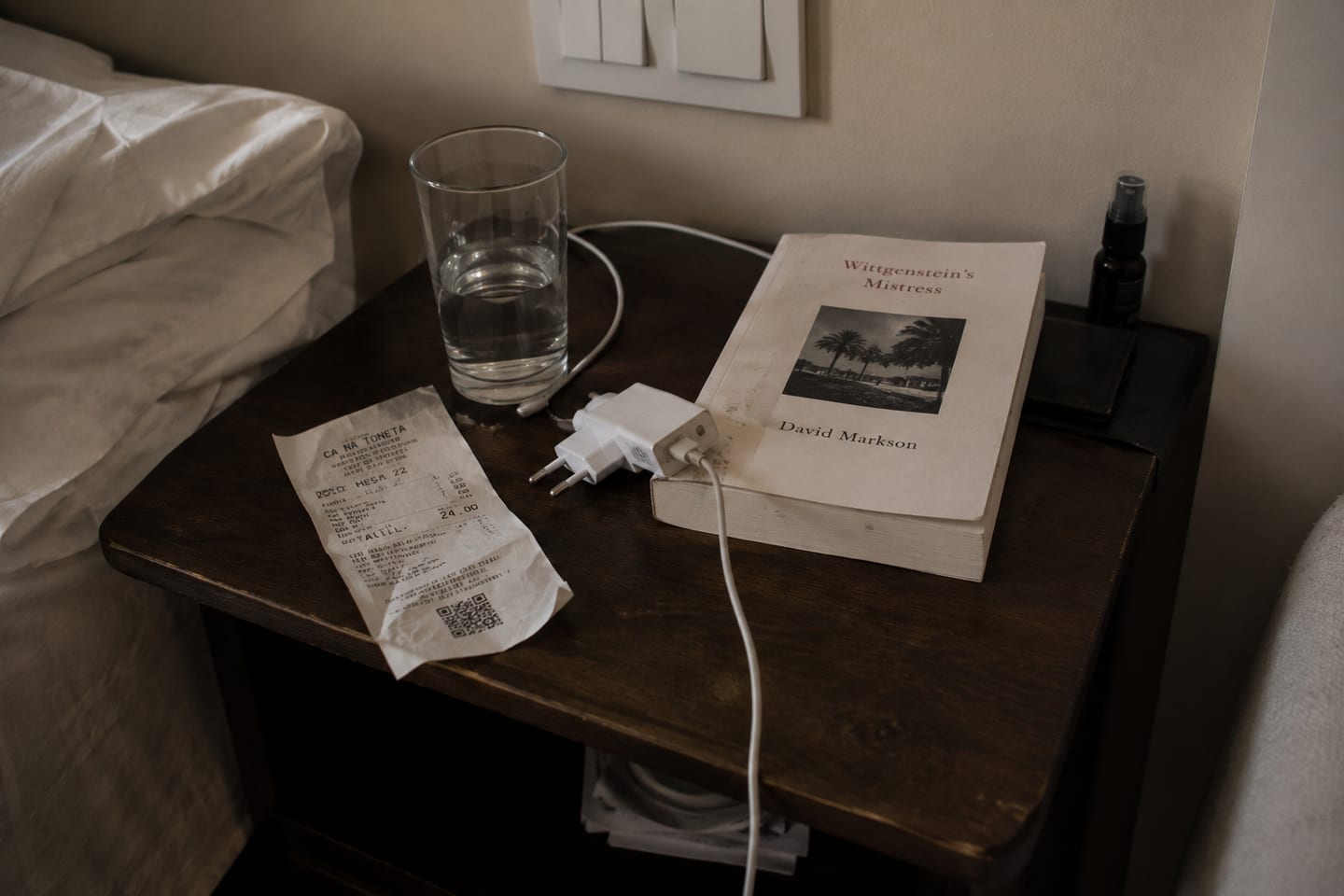 A dark wood bedside table — a glass of water, a folded paper receipt from Ca Na Toneta (€24, mesa 22), David Markson's *Wittgenstein's Mistress* with palms on the cover, a coiled white iPhone charger cable, rumpled white bed sheets at the left edge, low warm light.