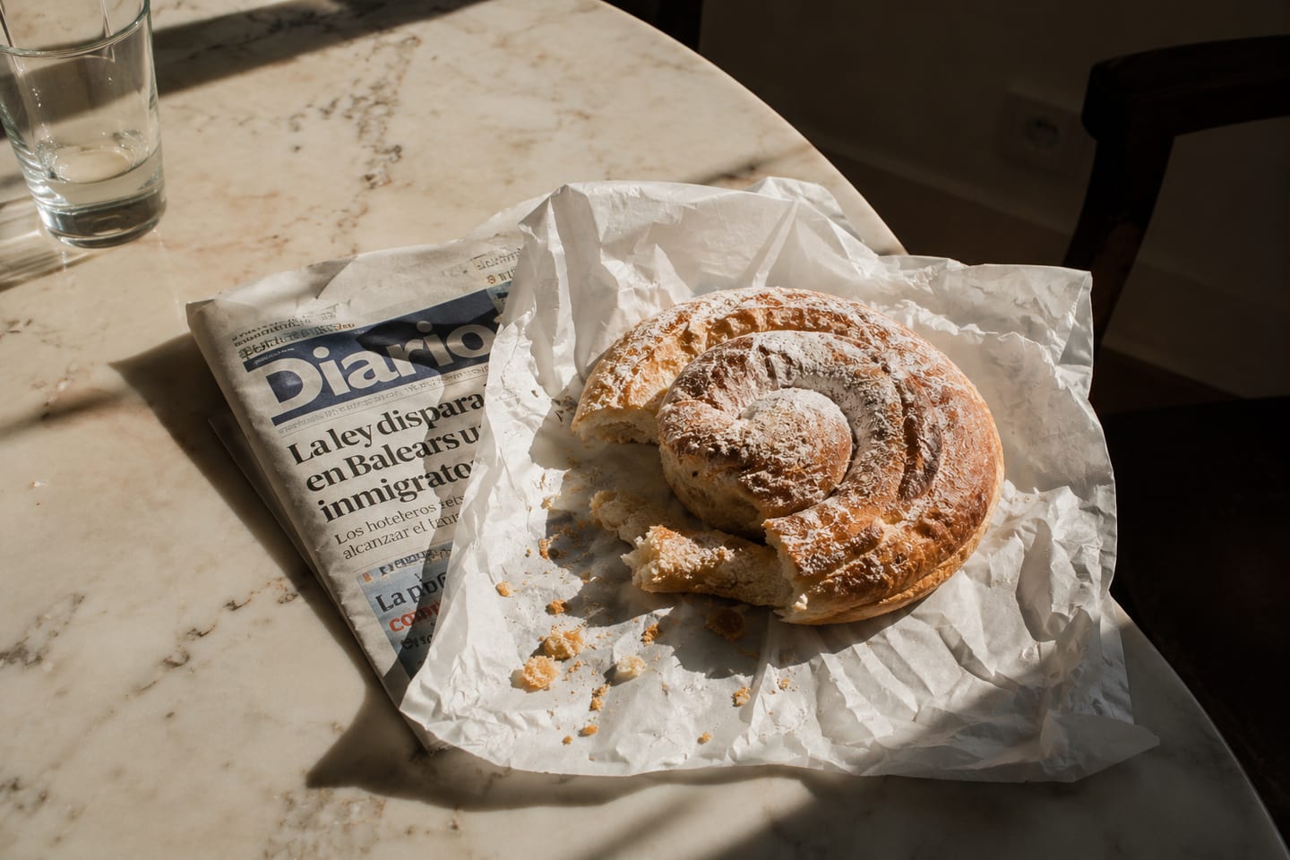 A round marble café table — a glass of water, a folded Diario de Mallorca, a torn ensaïmada on baker’s paper, slanted morning light.