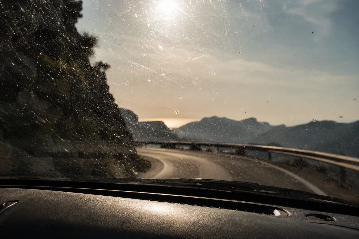 View through a dusty windshield with the dashboard in the foreground, a winding road curving right toward the Tramuntana ridge in the distance, sun catching the dust on the glass as a starburst, a tree silhouette at the left edge.