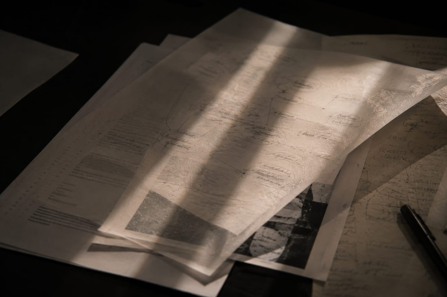 Translucent vellum tracing paper layered over typed papers and printed diagrams. Pencil flow-charts visible through the vellum, a small black-and-white architectural photograph peeking out, a black pen resting in the lower-right corner, light coming through shutter slats.