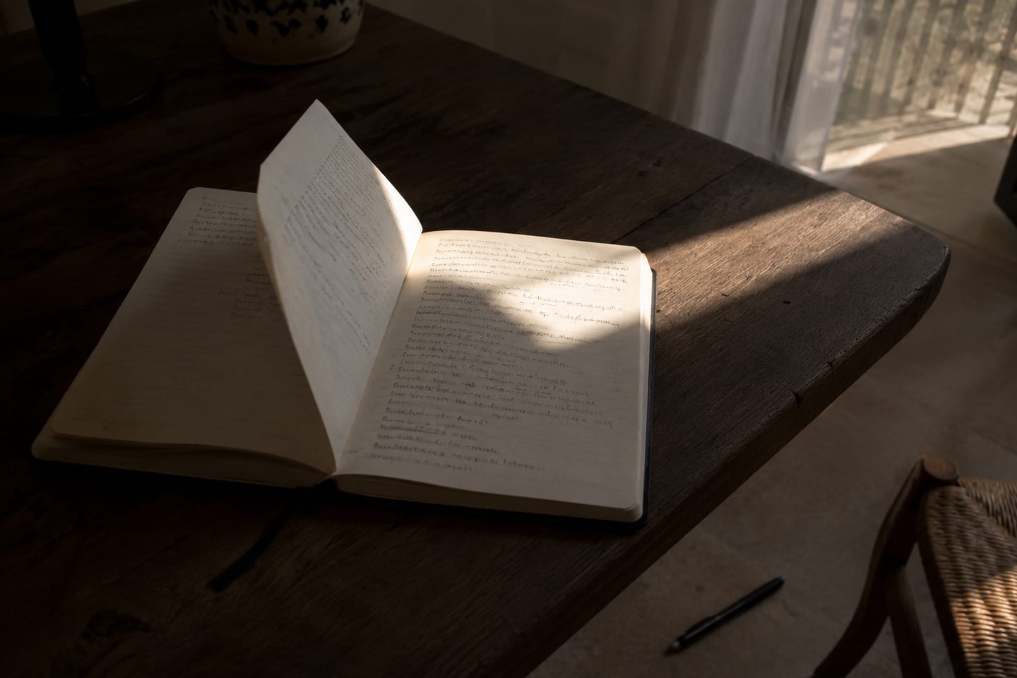 Open notebook on a dark wood table, a single diagonal shaft of warm afternoon light falling across the right page over handwritten cursive lines, a pen resting beside the spine, window soft beyond.