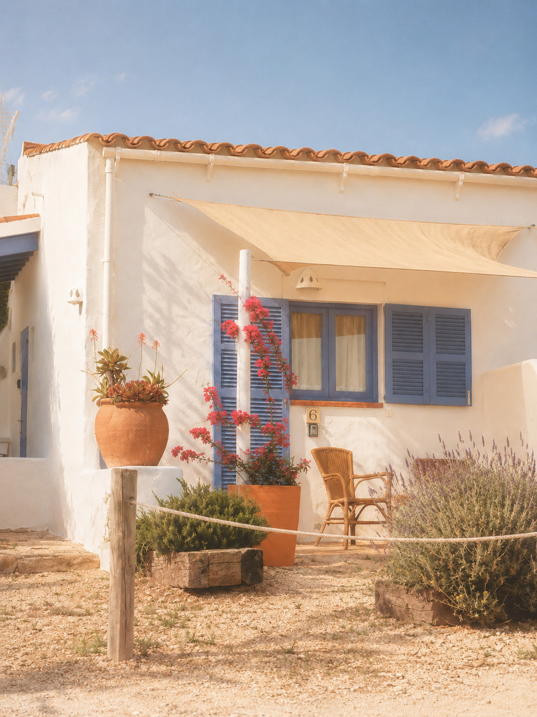 A whitewashed Formentera house with blue shutters, terracotta pots, bougainvillea and lavender, a wicker chair on a shaded terrace.