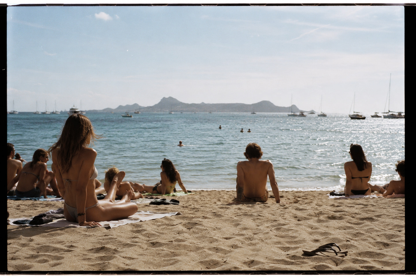 A Formentera beach in late afternoon, a row of bathers seated and lying along the sand, sea opening to a far island, the light low and warm.