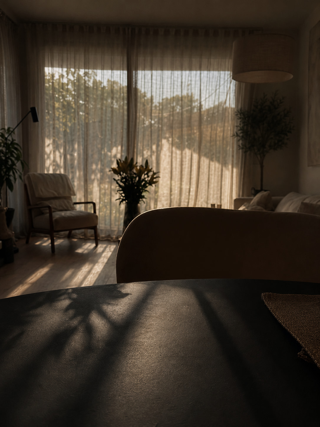 A wicker chair beside sheer curtains in golden afternoon light, flowers and a dark tabletop in the foreground.