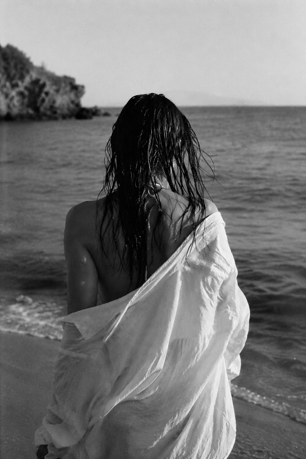 A woman from behind at the shoreline, wet hair, a white cloth wrapped loosely around her shoulders, the sea opening to a distant headland.