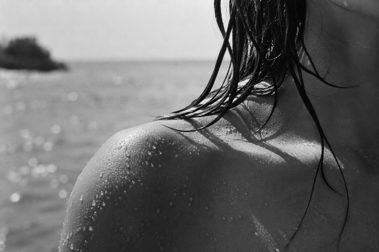 A bare shoulder in close-up, water droplets across the skin, wet hair falling, sea soft in the background.