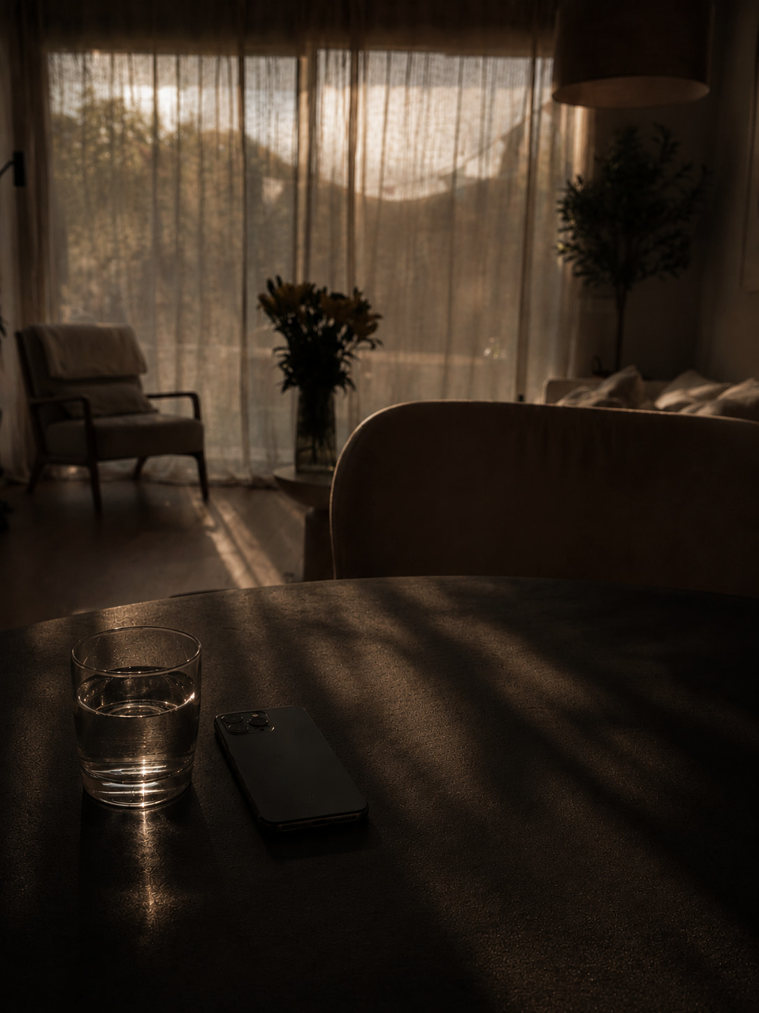 A dark wooden table in golden afternoon light, a glass of water and a phone resting on it, a chair and sheer curtains visible in the background interior.